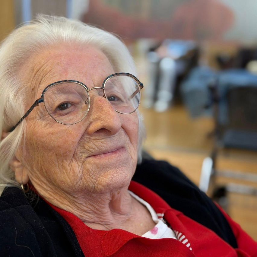 Eine ältere Frau mit weißen Haaren, die eine Brille und ein rotes Hemd trägt, posiert für ein Foto mit einem unscharfen Hintergrund aus Stühlen und Tischen.