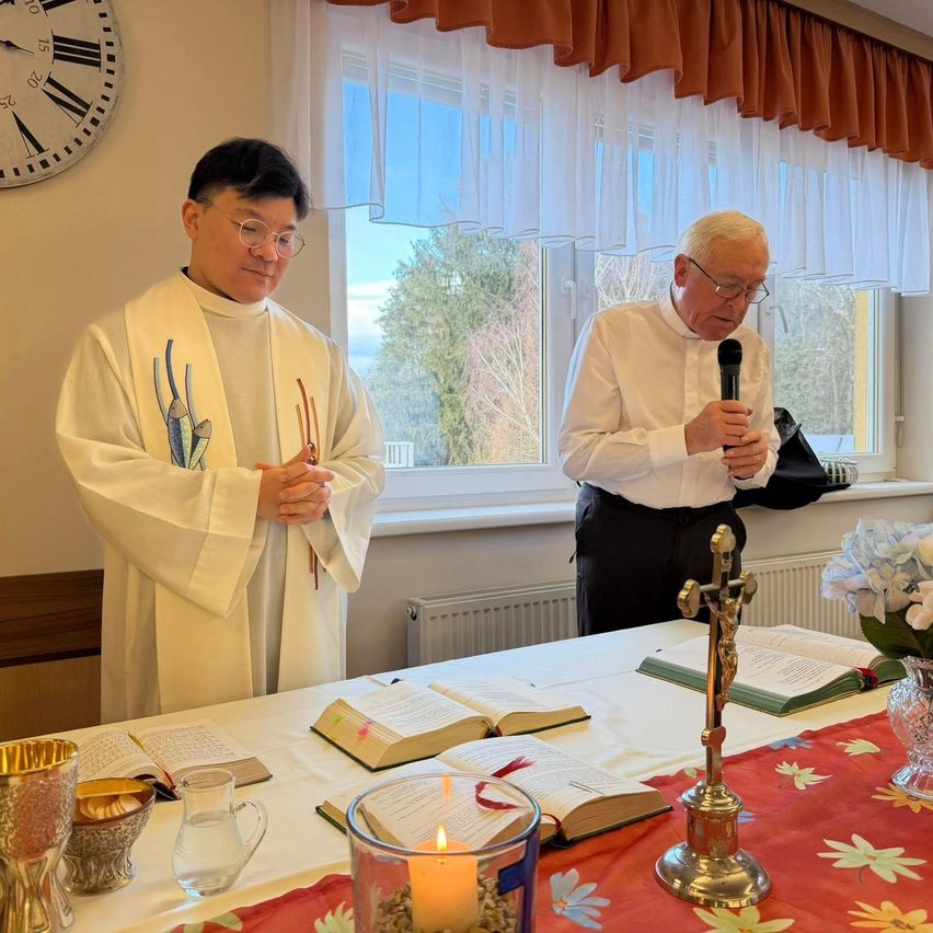 Zwei Priester stehen bei einer Zeremonie an einem Tisch, einer hält ein Mikrofon. Offene Bücher und eine Kerze liegen auf dem Tisch, und ein Kreuz steht zwischen ihnen. Eine Uhr und ein Fenster mit Vorhängen sind im Hintergrund.