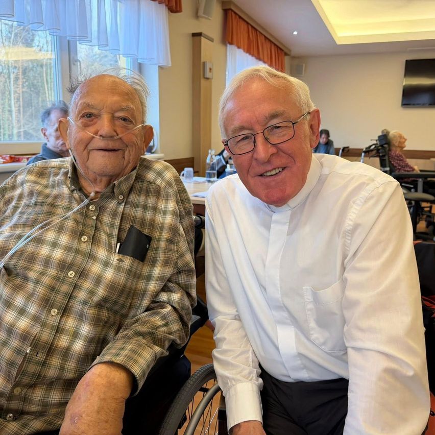 Zwei ältere Männer in Rollstühlen posieren für ein Foto in einem Raum. Der Mann auf der linken Seite trägt ein kariertes Hemd und eine Sauerstoffschlauch, während der Mann auf der rechten Seite ein weißes Hemd und eine Brille trägt. Hinter ihnen sitzen andere Leute auf Stühlen, ein Tisch mit Flaschen und ein Monitor.