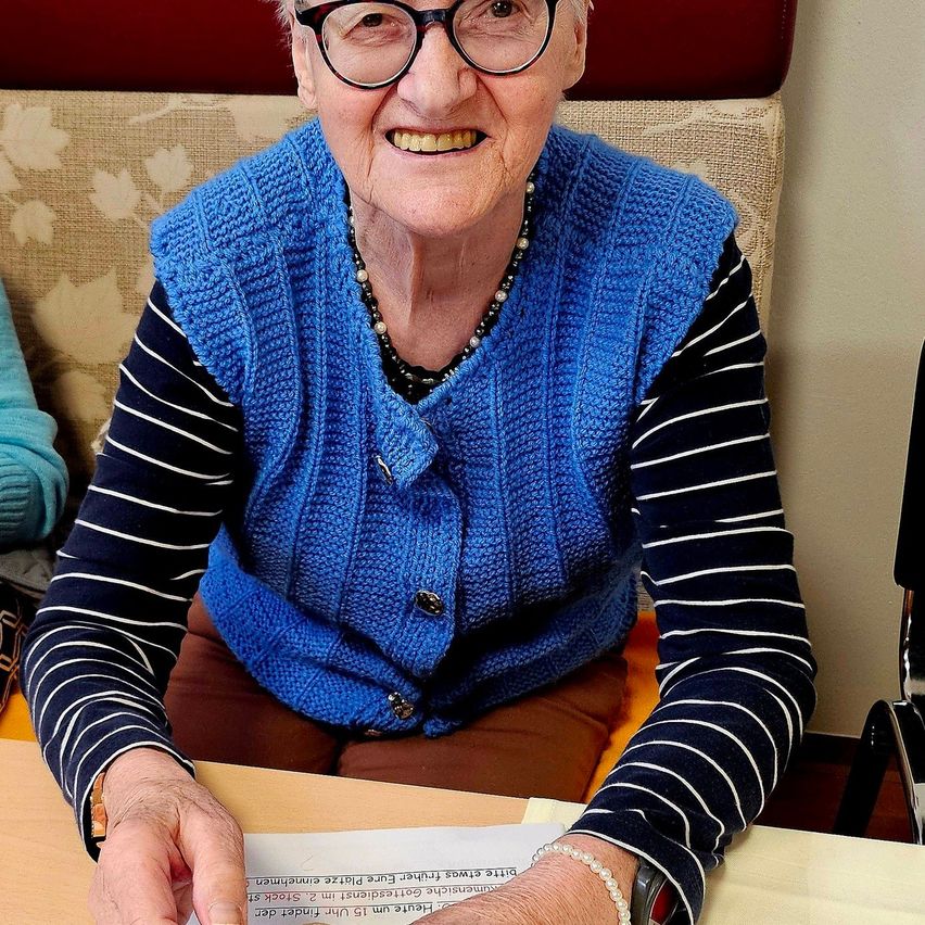 Eine ältere Frau mit Brille, einem blauen gestrickten Schal und einer Halskette sitzt in einem Stuhl und lächelt. Sie hält ein Stück Papier mit Text in der Hand.
