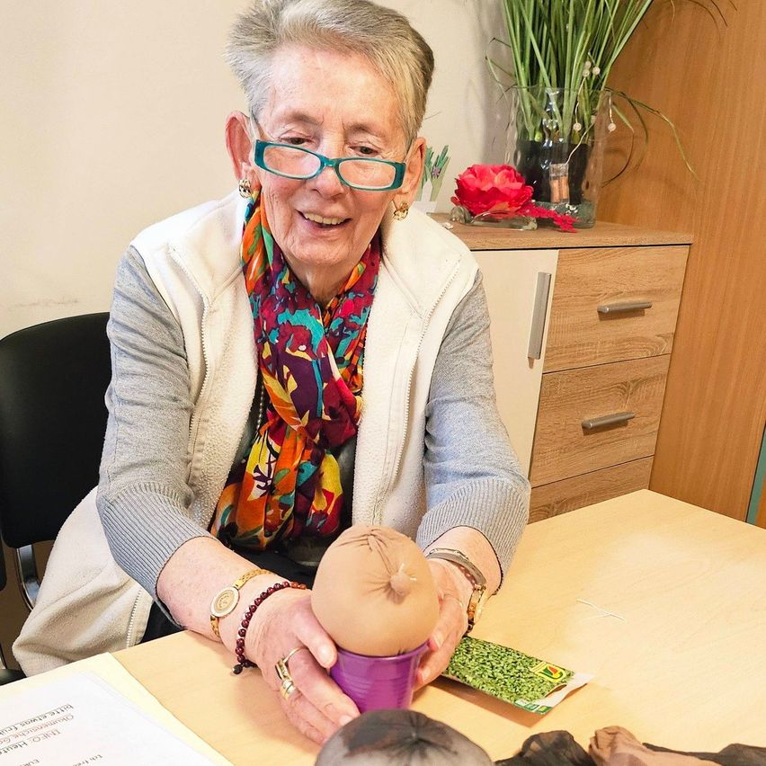 Eine ältere Frau mit Brille und einem Schal sitzt an einem Tisch und hält ein rosafarbenes Kopfmodell in der Hand und lächelt.