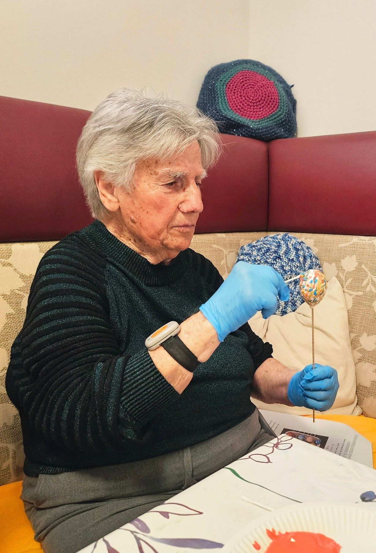Eine ältere Frau sitzt auf einem Stuhl. Sie trägt Handschuhe und dekoriert ein Osterei mit einem Stick. Sie hat einen ernsten Ausdruck im Gesicht.