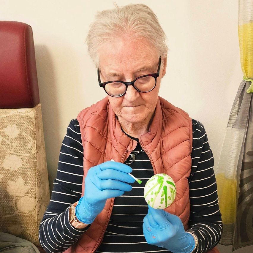 Eine ältere Frau mit Brille und blauen Handschuhen malt eine grüne Kugel mit einem Pinsel.
