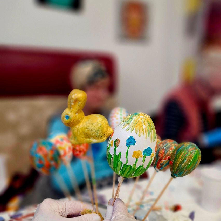 Eine Person hält ein dekoriertes Osterei in der Hand, um sie herum sind andere. Im Hintergrund sitzen zwei Personen mit verschwommenen Gesichtern.
