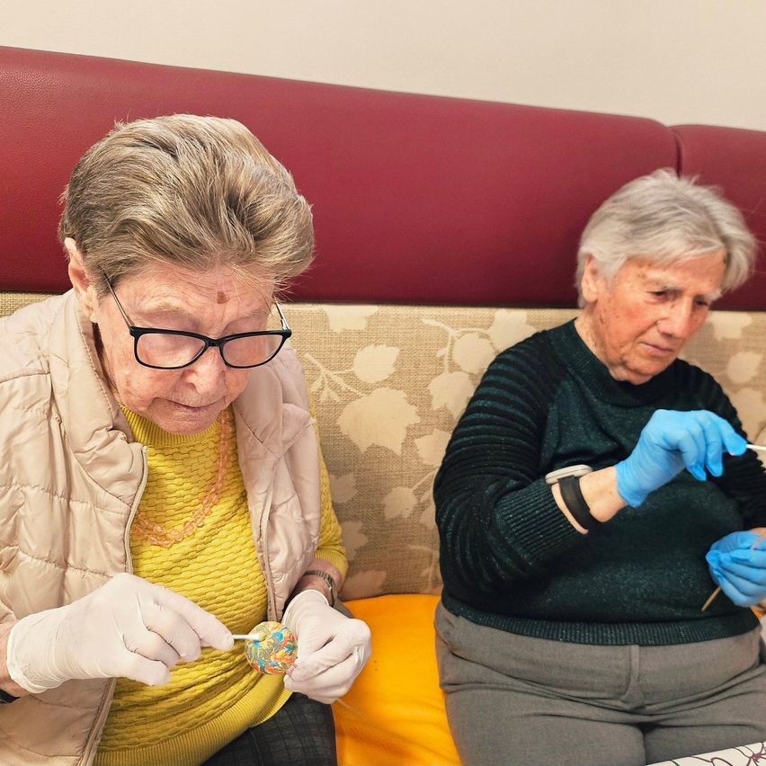 Zwei ältere Frauen, die Handschuhe tragen, sitzen auf einem Sofa. Eine bemalt ein Osterei mit einem Pinsel. Beide tragen eine Brille.