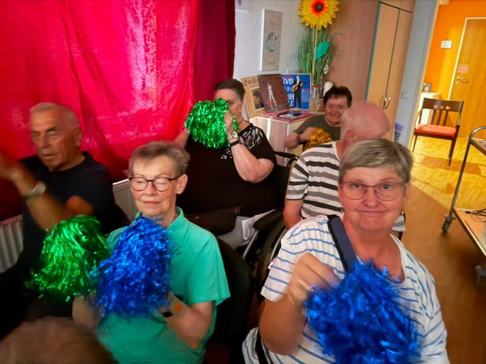 Eine Gruppe älterer Erwachsener in einem Raum, einige halten blaue und grüne Pompons und lächeln in die Kamera. Ein roter Vorhang und eine gelbe Blume sind im Hintergrund.