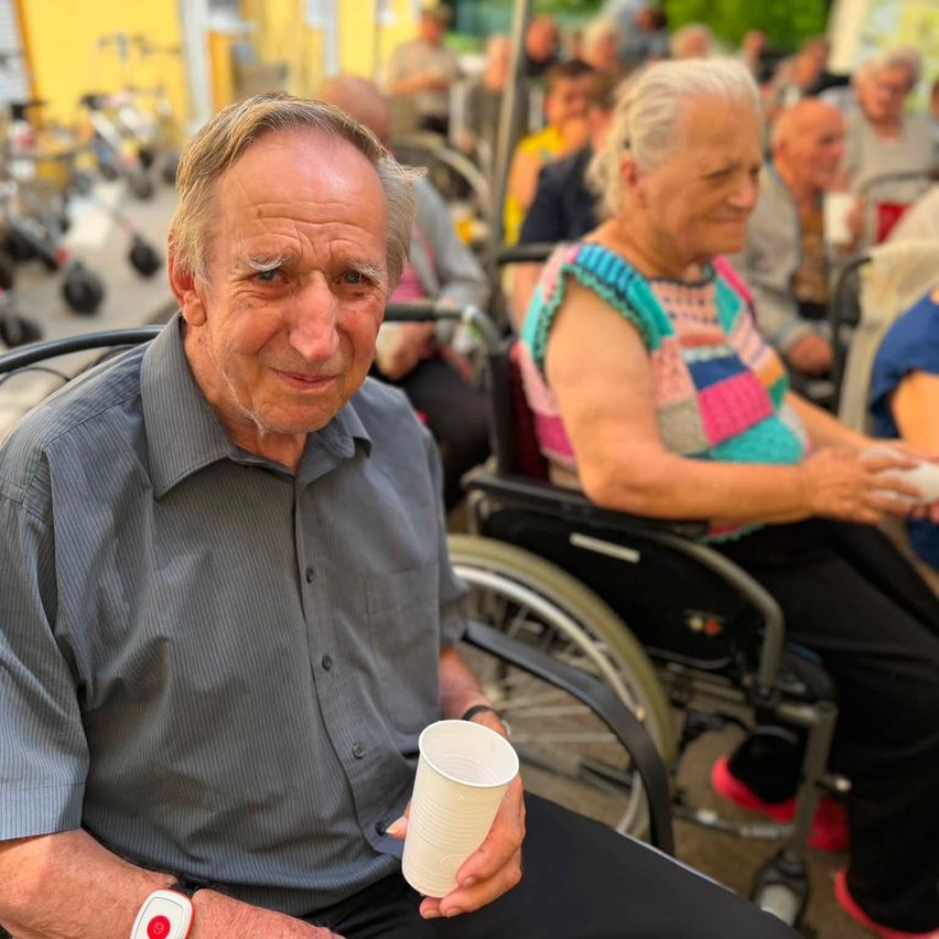 Ein älterer Mann im Rollstuhl lächelt und hält einen weißen Becher, während eine Frau in einem bunten Oberteil hinter ihm im Rollstuhl sitzt.