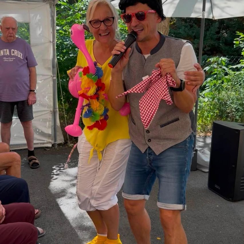 Ein Mann und eine Frau stehen zusammen, der Mann hält ein Mikrofon und einen rosa Flamingo, die Frau hält einen rosa Flamingo und eine bunte Girlande. Sie lächeln und scheinen bei einer Veranstaltung im Freien aufzutreten.