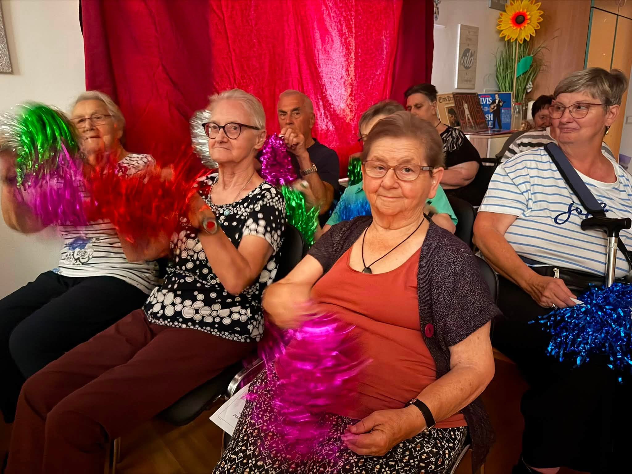 Seniorengruppe mit Pompons in bunten Farben in einem Raum mit rotem Vorhang.