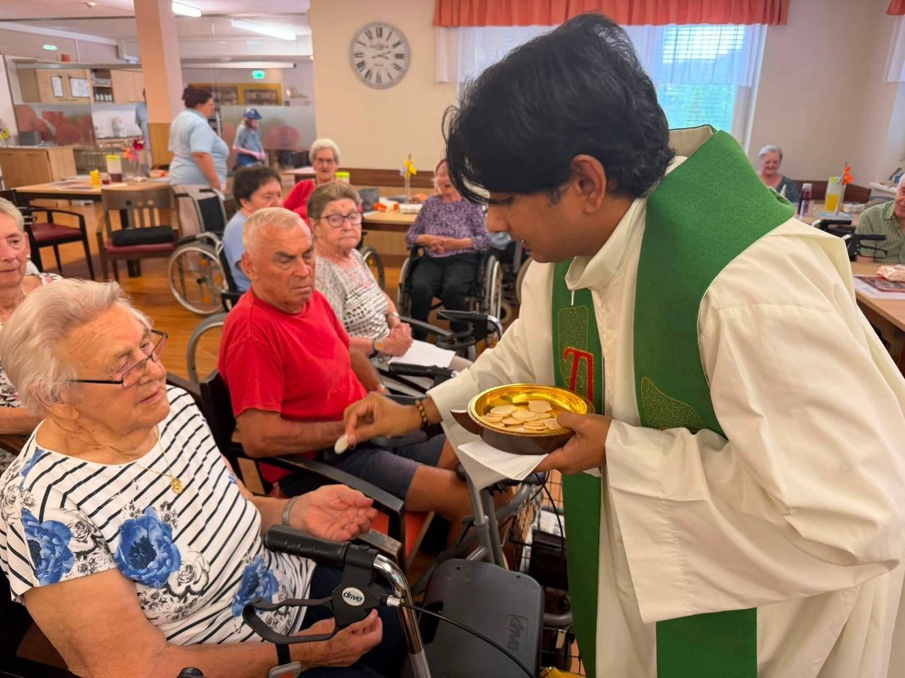 A priest in a green and white robe is giving communion to an elderly woman in a wheelchair.