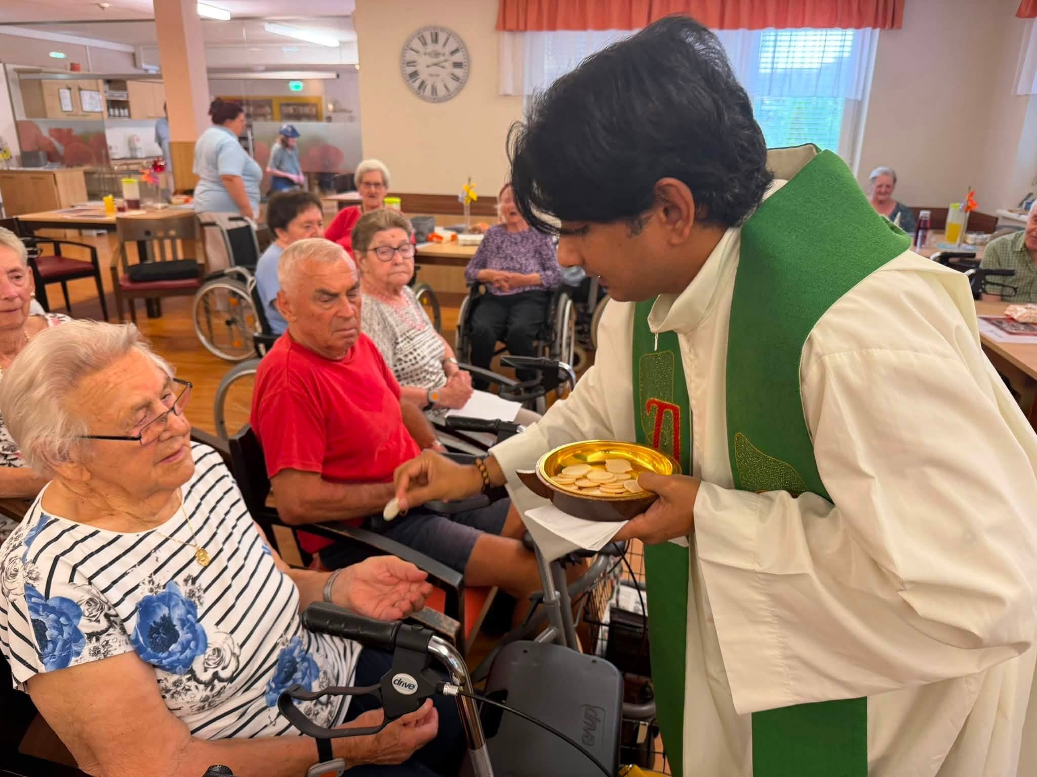 Priester teilt Kommunion mit Senioren in einem Gemeinschaftsraum in Österreich