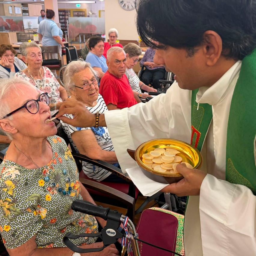 A priest feeds a communion wafer to an elderly woman in a wheelchair, surrounded by other elderly individuals in a room.
