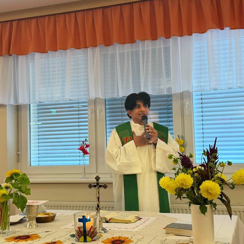 A priest stands in front of a table with flowers and a candle, holding a microphone, in a room with white curtains and a window.
