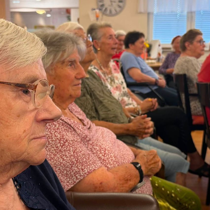 A group of elderly women sitting in chairs, possibly in a meeting or social gathering. They are smiling and appear to be engaged in conversation. The room has a warm, cozy atmosphere.