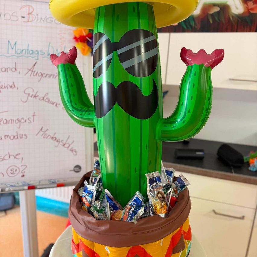 A large inflatable cactus with a yellow hat, sunglasses, and a mustache stands in front of a whiteboard. It's filled with candy and placed on a table.
