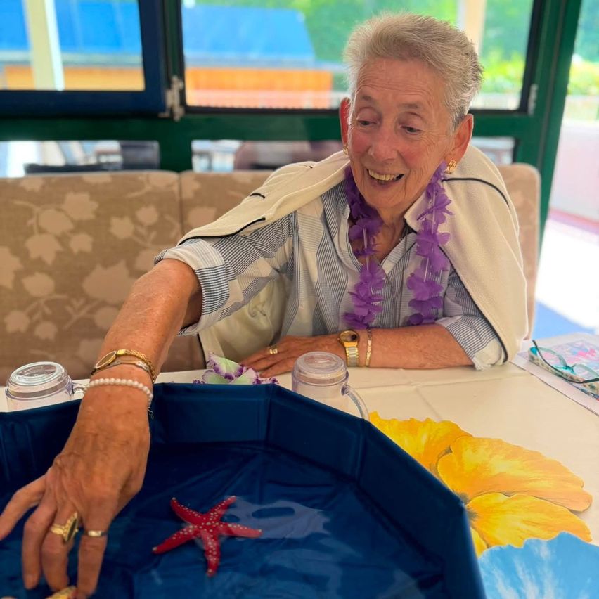 An elderly woman wearing a purple lei smiles while holding a red starfish in a blue tray with a floral design on the table.