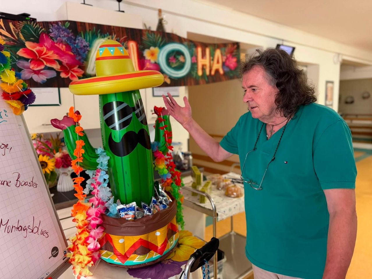 An older man with glasses on a stand speaks to a large inflatable cactus with a sombrero and mustache. Behind him is a table with food and a banner with the word 'O HA'.
