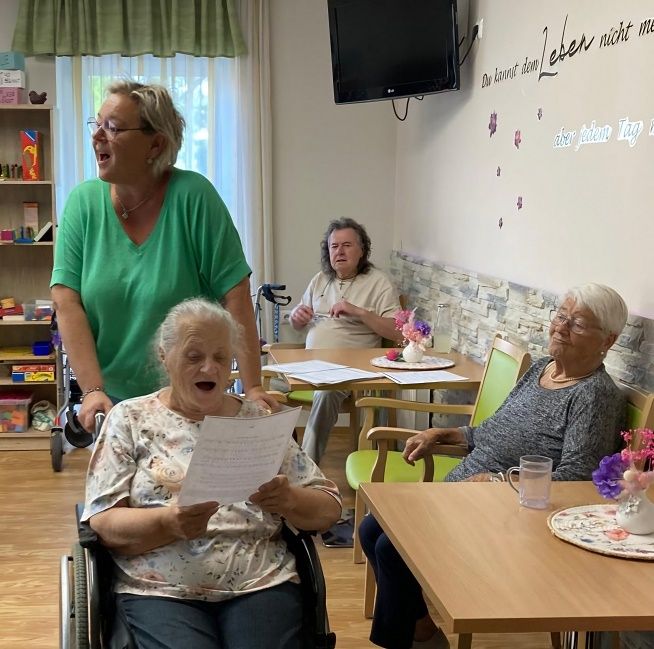 Eine Gruppe älterer Frauen in einem Raum, eine liest ein Papier, eine andere lacht und eine dritte sitzt mit Blumen auf dem Tisch. Ein Fernseher ist an der Wand montiert.
