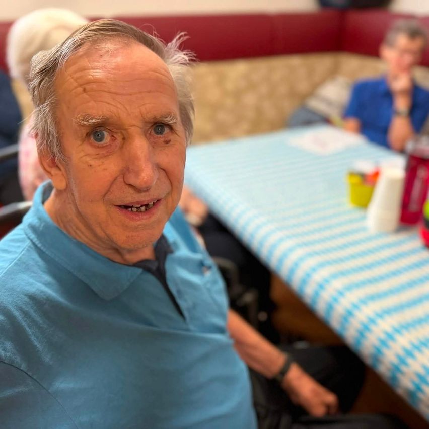 Ein älterer Mann mit grauem Haar und einem blauen Poloshirt lächelt in einer sozialen Umgebung, mit einem Tisch und anderen Menschen im Hintergrund.