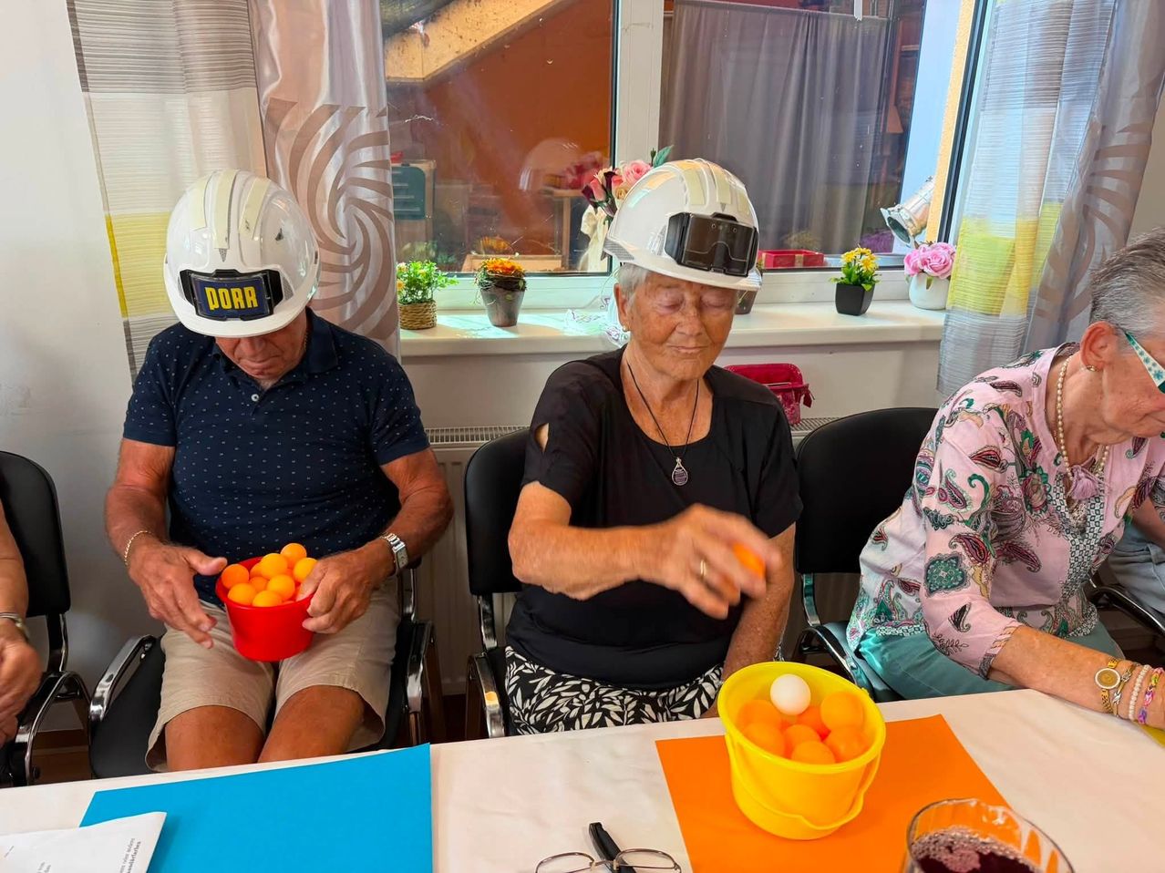 Zwei ältere Personen mit weißen Schutzhelmen sitzen an einem Tisch, eine hält einen roten Becher mit Tischtennisbällen, während die andere versucht, einen Ball mit einem Schläger zu treffen.