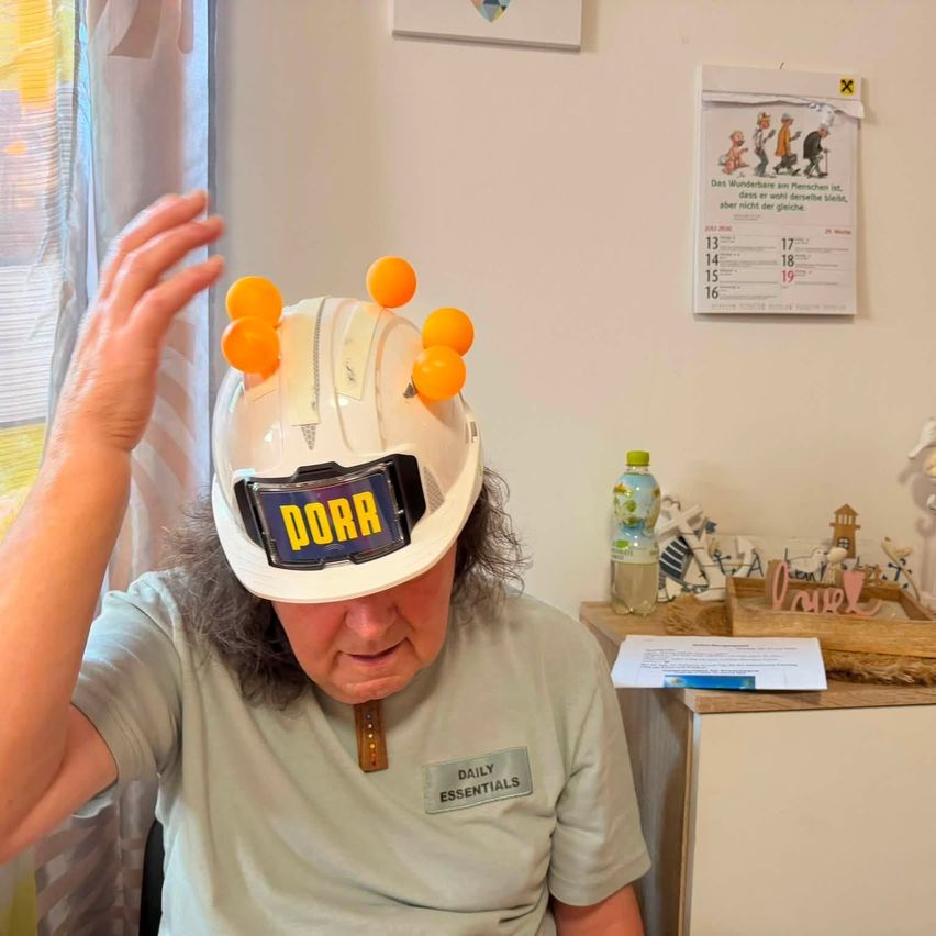 Eine ältere Frau trägt einen weißen Schutzhelm mit orangefarbenen Kugeln auf der Oberseite und einem Namensschild mit der Aufschrift 'Daily Essentials'. Sie befindet sich in einem Raum mit einem Kalender an der Wand und einem Schreibtisch mit verschiedenen Gegenständen.