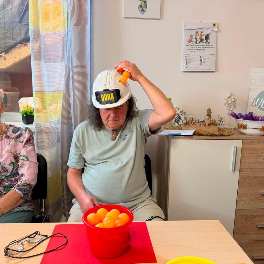 Ein Mann mit einem weißen Helm mit dem Wort DORA sitzt an einem Tisch mit einer Schüssel Orangen. Eine Frau sitzt neben ihm.