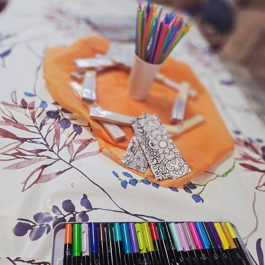 Ein Tisch mit einem floralen Tischtuch zeigt eine Sammlung von bunten Markern, einem kleinen orangefarbenen Tuch und einem weißen Becher gefüllt mit Strohhalmen.