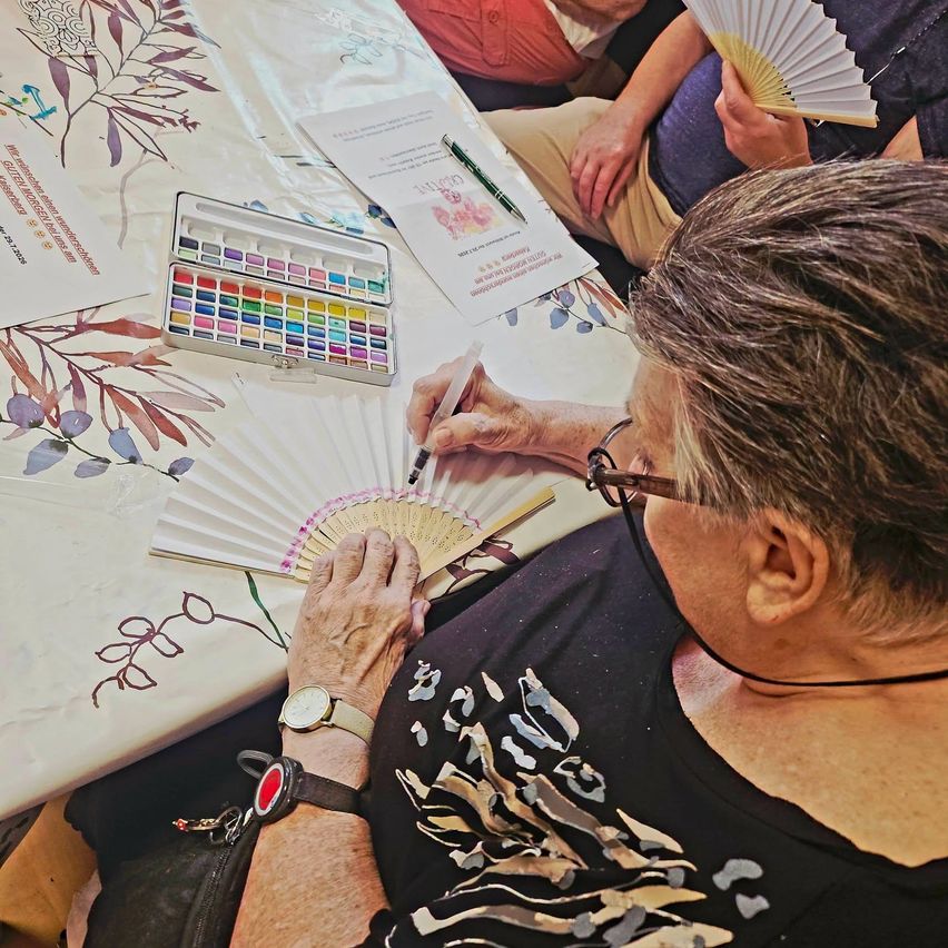 Eine ältere Frau malt ein Blumendesign auf einen Papierfächer mit Aquarellfarben. Vor ihr steht ein Tisch mit einer blumigen Tischdecke.