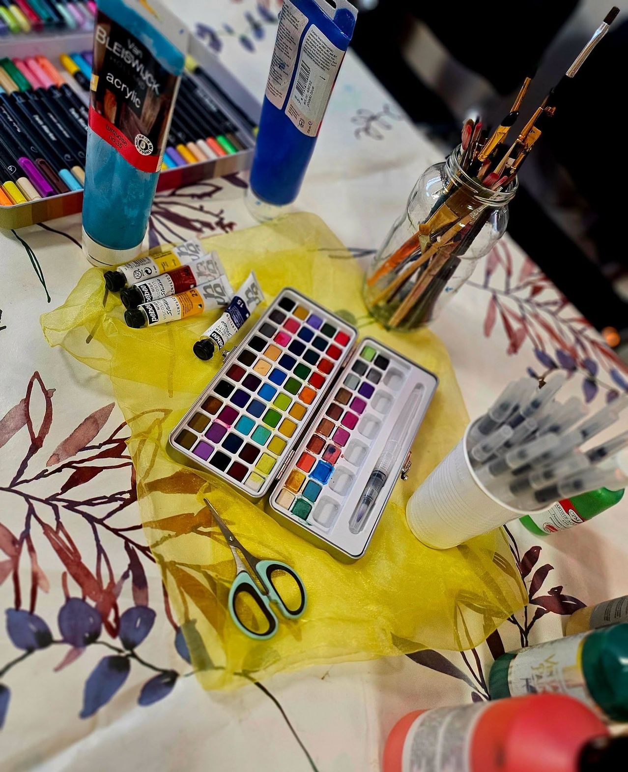 Ein Tisch mit verschiedenen Künstlerbedarfsartikeln, einschließlich eines Satzes Aquarellfarben, Pinsel und Stifte. Das Aquarellset ist geöffnet, und Scheren liegen auf einem gelben Tuch.