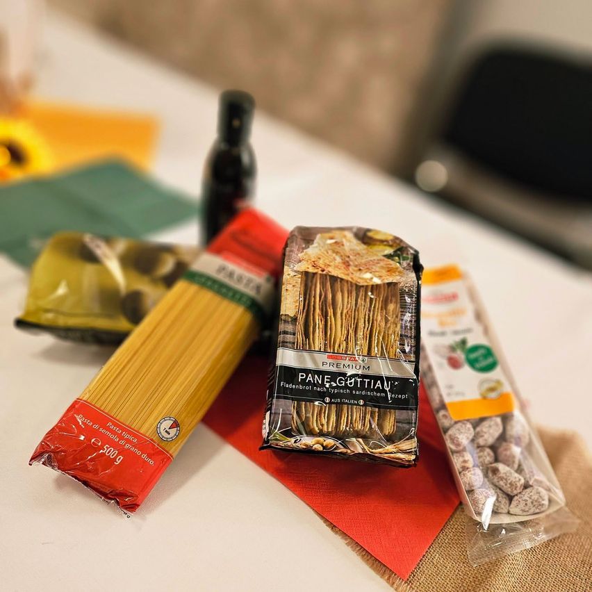 Auf einem Tisch befinden sich eine Flasche Sauce, eine Tüte Nudeln und eine Tüte Nüsse. Die Nudeln sind mit 'Pane e Guttian' beschriftet und die Nüsse sind in einer durchsichtigen Packung.