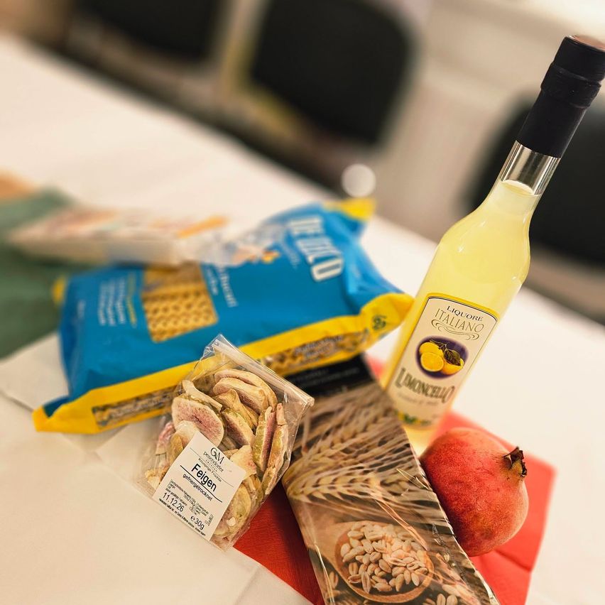 Auf einem Tisch stehen eine Flasche Limoncello, eine Tüte Feigen, eine Tüte Weizen und ein Granatapfel.
