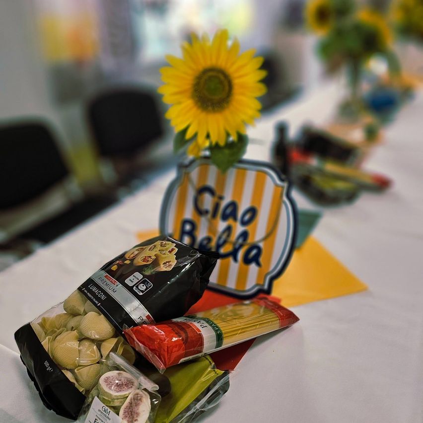Ein Tisch mit Sonnenblumen-Zentrierung, einem 'Ciao Bella'-Schild und verschiedenen Lebensmitteln wie Nudeln, Feigen und Nudelstange.