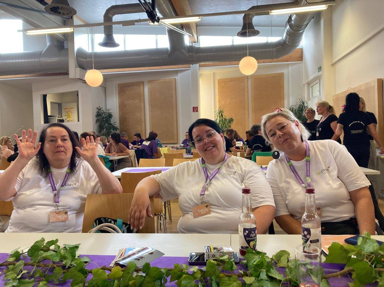 Three women wearing white shirts with purple lanyards sit at a table in a room. They are surrounded by bottles and greenery. Other people sit at other tables in the background.