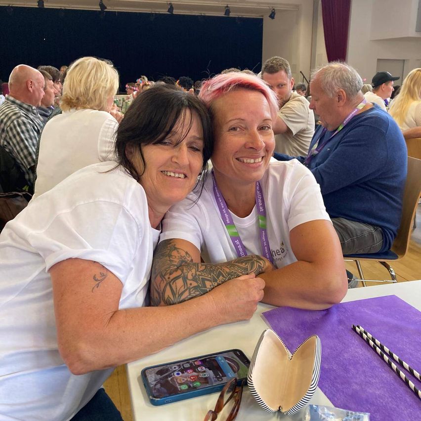 Two women smile for a picture at an event. They are sitting at a table with a cell phone and glasses case on it. Other people sit behind them.