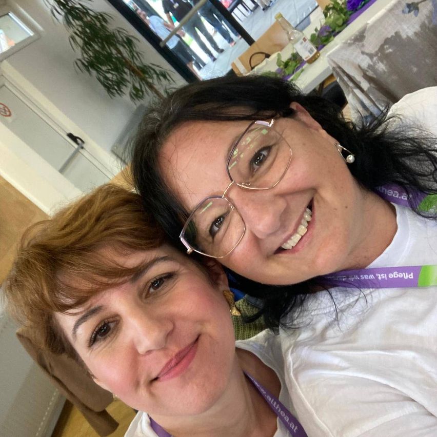 Two women are smiling for a photo. One is wearing glasses and a purple lanyard. The other is wearing a white shirt and has short hair. Behind them is a glass door with a plant and a table.