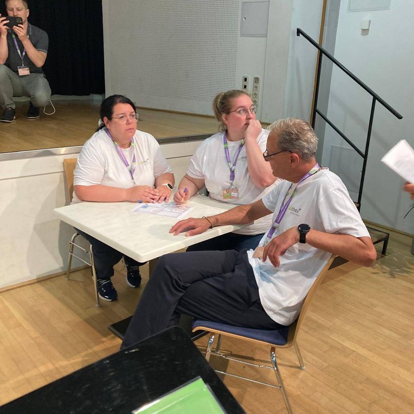 Three people are seated at a table, reviewing documents. One person is writing, while the others are observing. A man takes a photo from behind.