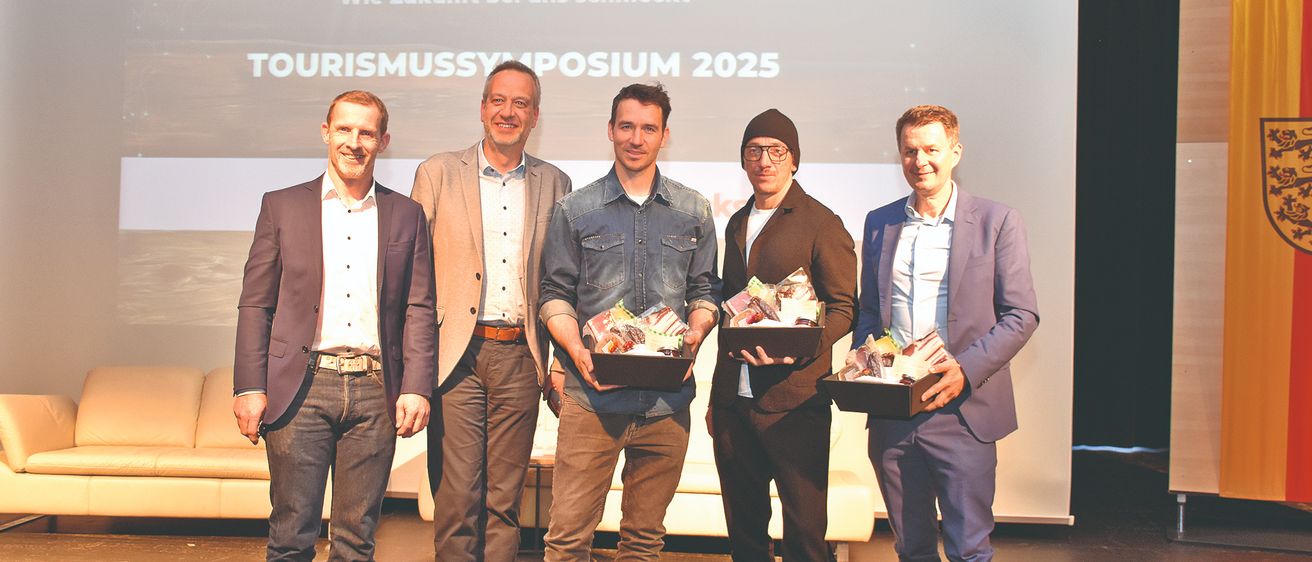 Fünf Männer stehen auf einer Bühne und halten Boxen mit Essen. Hinter ihnen befindet sich ein Bildschirm mit dem Text 'Klund Kaiseam Wie Zukunft bei uns schmeckt' und 'TOURISMUSSYMPOSIUM 2025'. Sie scheinen sich auf einer Veranstaltung zu befinden.