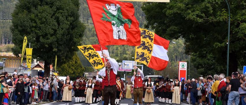 Eine Gruppe von Männern in traditionellen Kostümen hält Flaggen in einer Parade. Ein Banner mit der Aufschrift 'Käsefestival' hängt über ihnen. Menschen säumen die Straße und schauen zu.