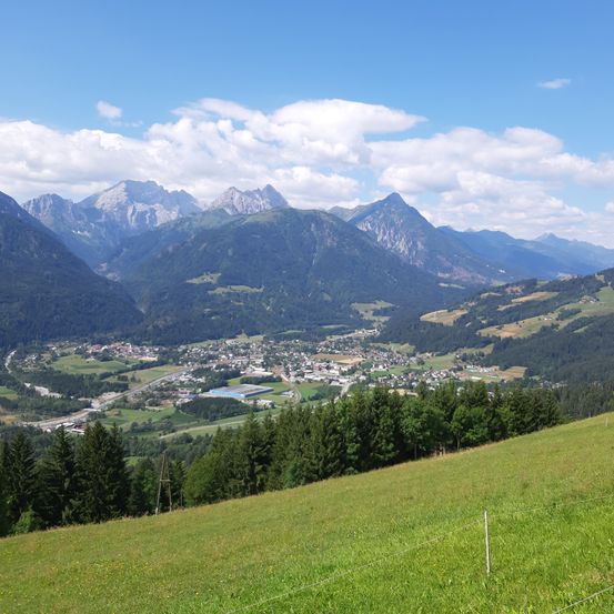 Eine Berglandschaft zeigt eine Stadt, eingebettet in üppige grüne Täler und dichte Wälder unter einem blauen Himmel mit verstreuten Wolken.