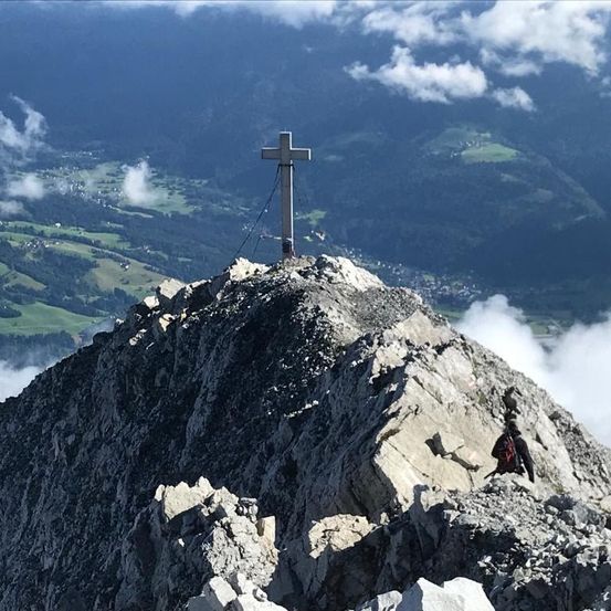 Luftaufnahme eines Berggipfels mit einem großen Kreuz auf seinem Gipfel, umgeben von felsigem Gelände und einem entfernten Tal mit verstreuten Gebäuden.