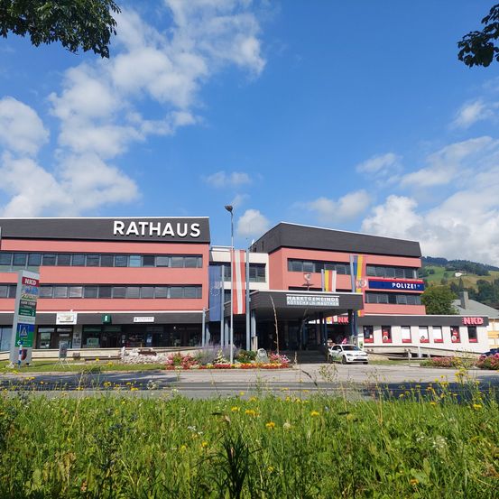 Das Rathaus-Gebäude ist in einer Stadt prominent zu sehen. Es ist rot gestrichen mit weißen Fenstern. Der blaue Himmel mit weißen Wolken ist oben sichtbar.