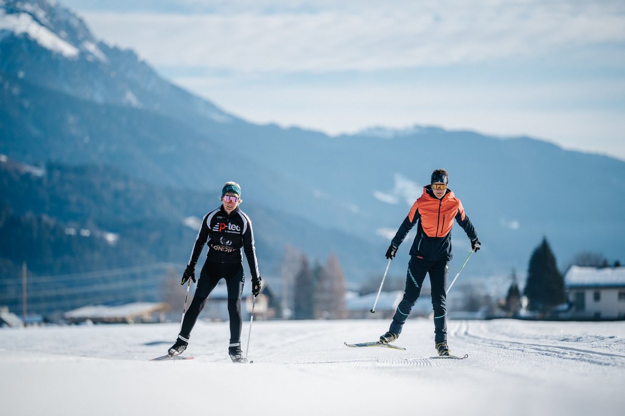 Zwei Skifahrer in Sportbekleidung sind auf einem verschneiten Hang im Langlauf unterwegs, mit Bergen im Hintergrund. Der Skifahrer auf der linken Seite trägt eine Schutzbrille und eine weiße und schwarze Kleidung. Der Skifahrer auf der rechten Seite trägt eine orange und schwarze Kleidung.