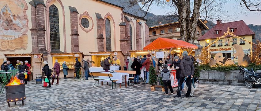 Ein überfüllter Außenmarkt in der Nähe einer Kirche mit verschiedenen Ständen und einkaufenden Menschen, einschließlich eines Kindes mit einem Hund an der Leine.
