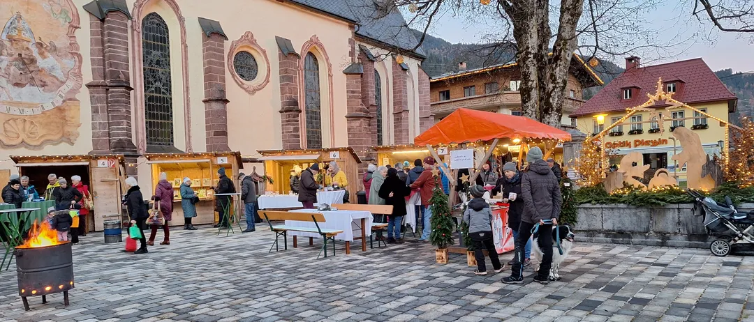 Ein überfüllter Außenmarkt in der Nähe einer Kirche mit verschiedenen Ständen und einkaufenden Menschen, einschließlich eines Kindes mit einem Hund an der Leine.