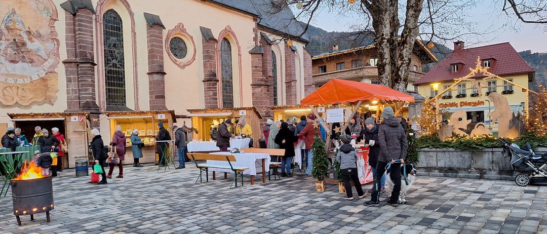 Ein überfüllter Außenmarkt in der Nähe einer Kirche mit verschiedenen Ständen und einkaufenden Menschen, einschließlich eines Kindes mit einem Hund an der Leine.