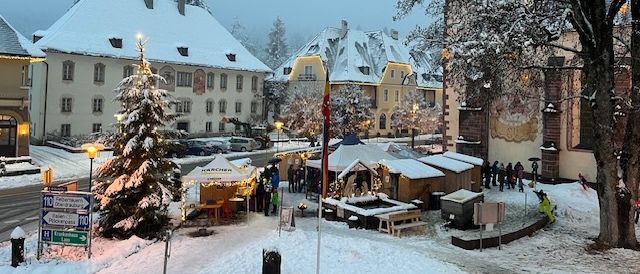 Ein verschneiter Stadtplatz mit einem Weihnachtsbaum, Marktständen und Menschen, die herumlaufen. Die Gebäude sind schneebedeckt, und es gibt Straßenlaternen und Bäume in der Umgebung.