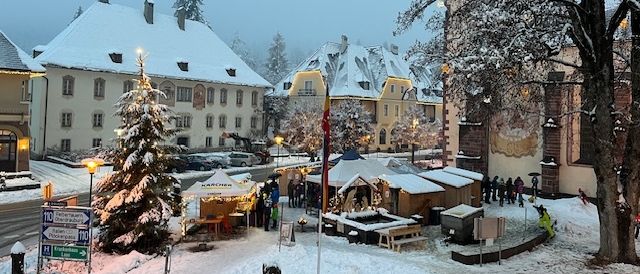 Ein verschneiter Dorfplatz mit einem Weihnachtsbaum und festlichen Ständen. Menschen gehen herum, und es gibt Gebäude mit schneebedeckten Dächern.