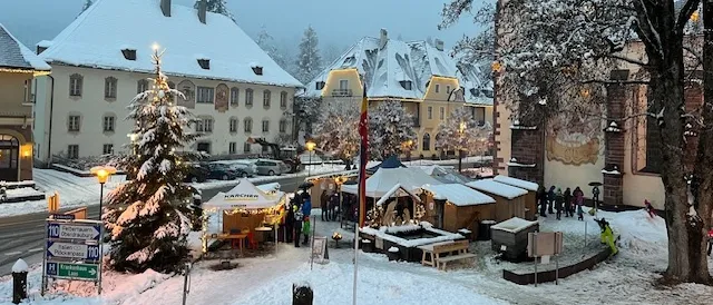 Ein verschneiter Stadtplatz mit einem Weihnachtsbaum, festlichen Ständen und umhergehenden Menschen. Gebäude sind verschneit, und Straßenlaternen sind eingeschaltet.