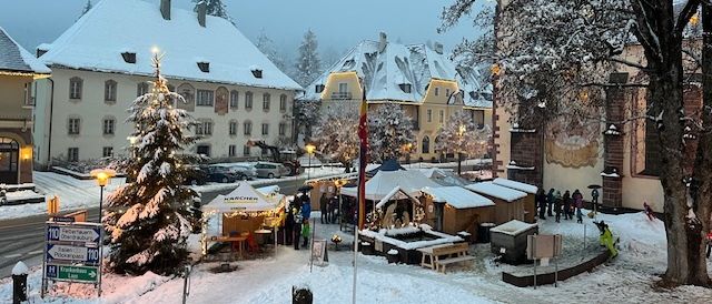 Ein verschneiter Stadtplatz mit einem geschmückten Weihnachtsbaum, kleinen Marktständen und Menschen, die herumlaufen. Die Gebäude sind schneebedeckt.