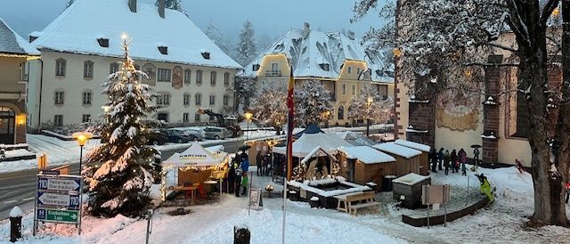 Ein verschneiter Stadtplatz mit einem Weihnachtsbaum, Marktständen und Menschen, die herumlaufen. Die Gebäude sind schneebedeckt, und die Straßenlaternen sind beleuchtet.